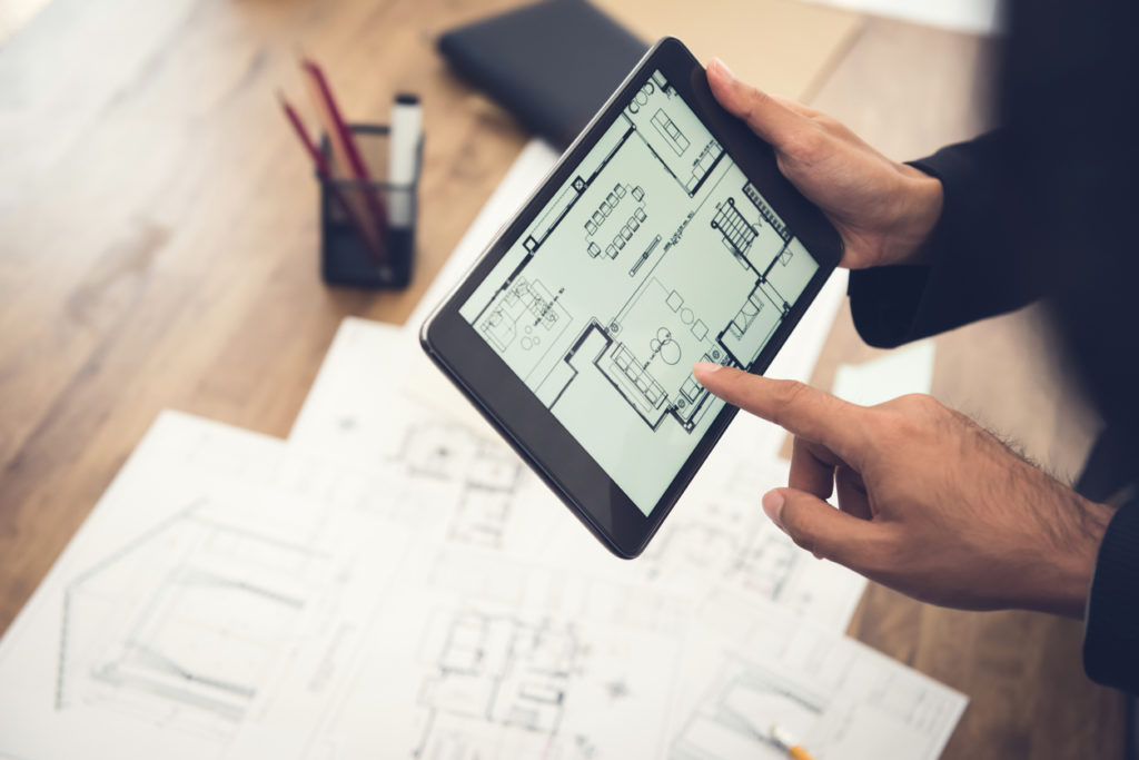 Architects holding a tablet device with a scanned drawing plan, showing architectural blueprints in the background.
